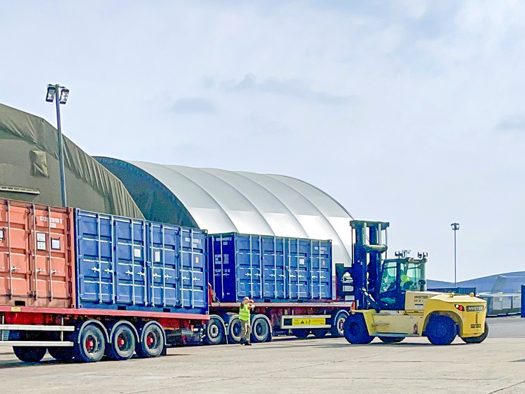 A yellow Hyster heavy-duty forklift stands next to a flatbed trailer loaded with blue shipping containers outside a large arched hangar.